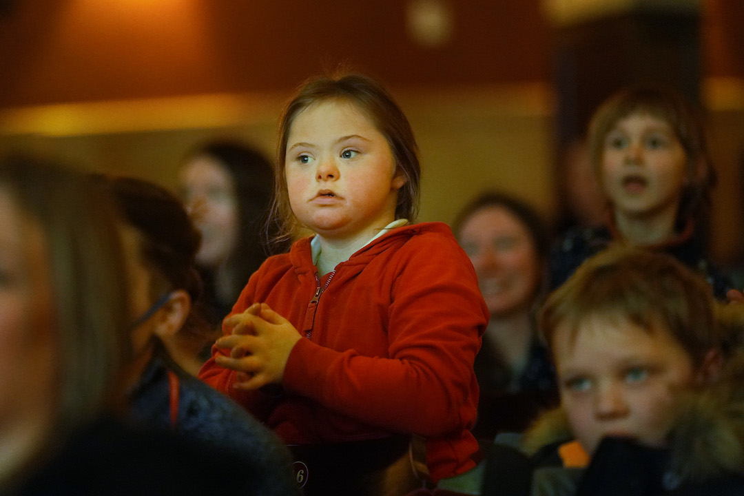 A young girl in a red hoodie sits among a group of people, appearing attentive and focused on something off-camera.