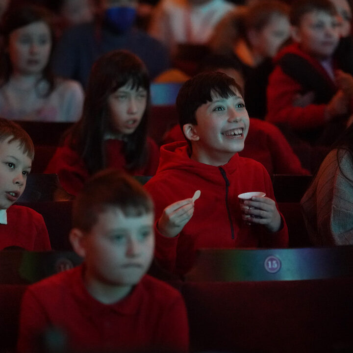 A group of children in red clothing sit in theatre seats, watching something on stage with various facial expressions of interest and excitement.