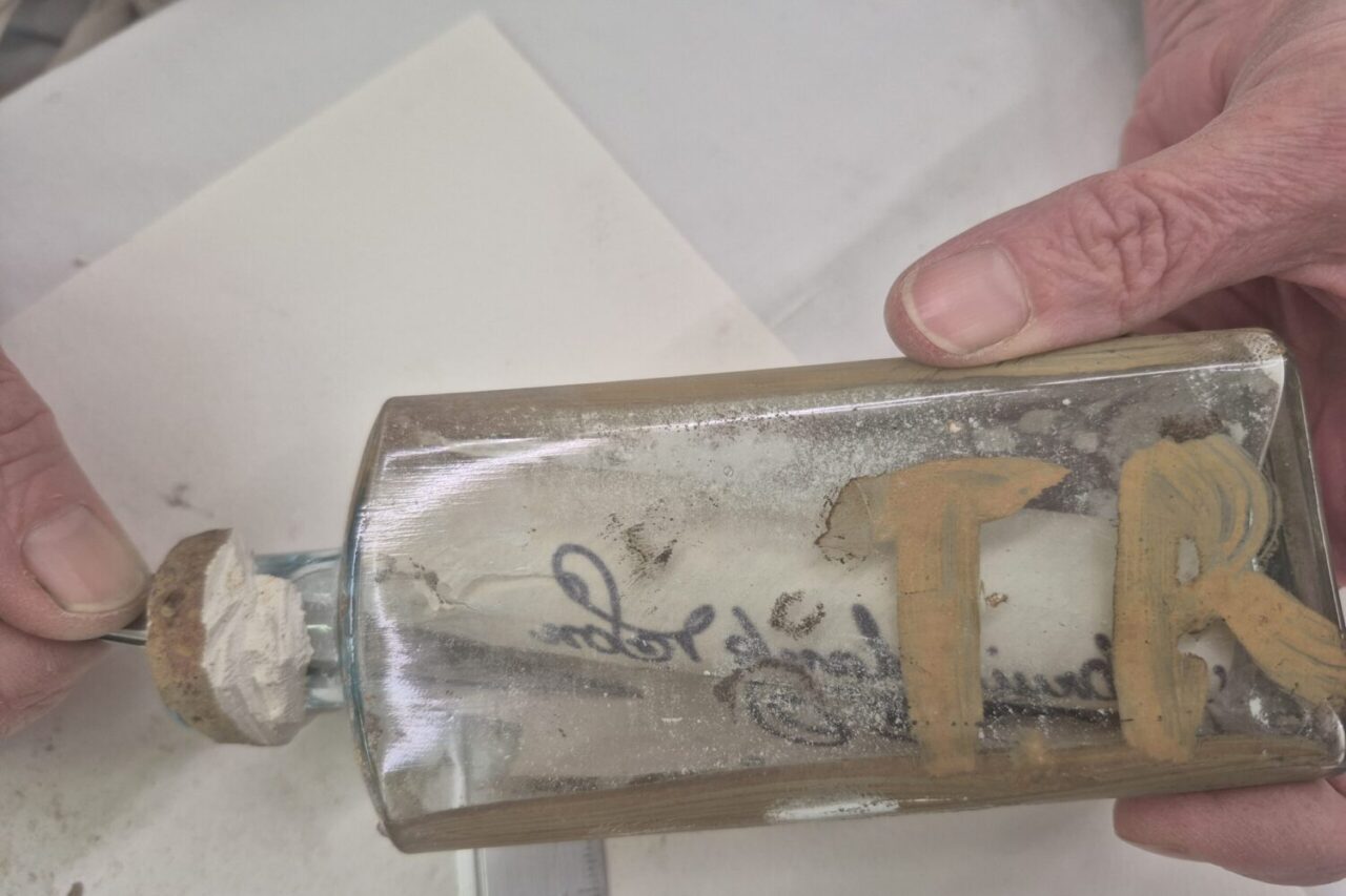 A close-up of hands holding an old, dusty glass bottle with a plaster stopper, a message inside and brown initials on the side that are difficult to read.