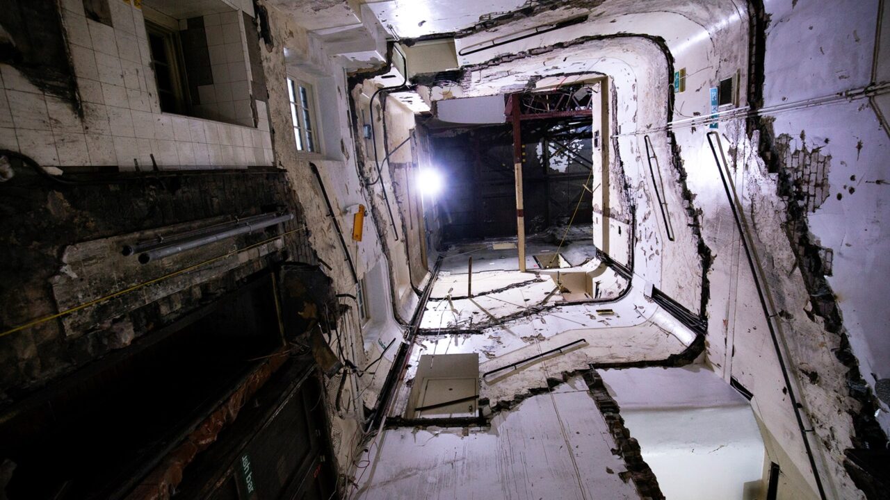 View looking up through the interior of a dilapidated, multi-story building with exposed walls, wires, and structural damage.