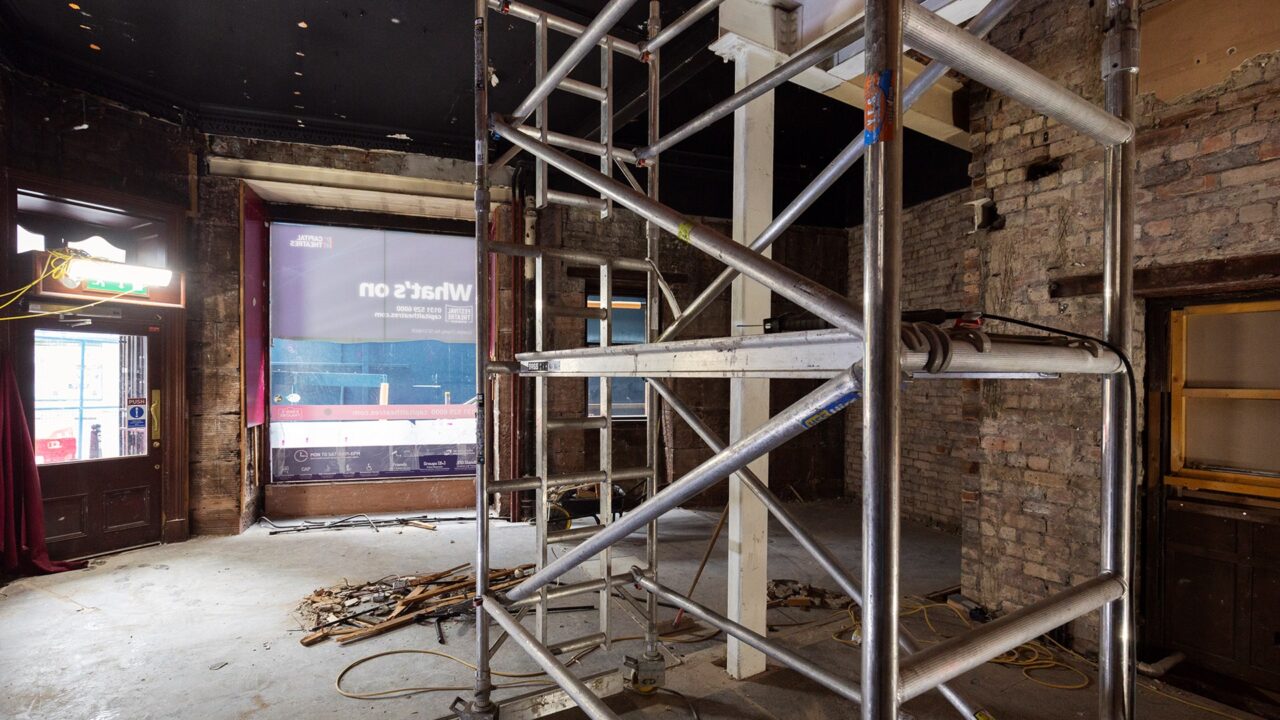 Interior of a building under renovation with scaffolding, exposed brick walls, construction materials, and tools scattered on the floor.