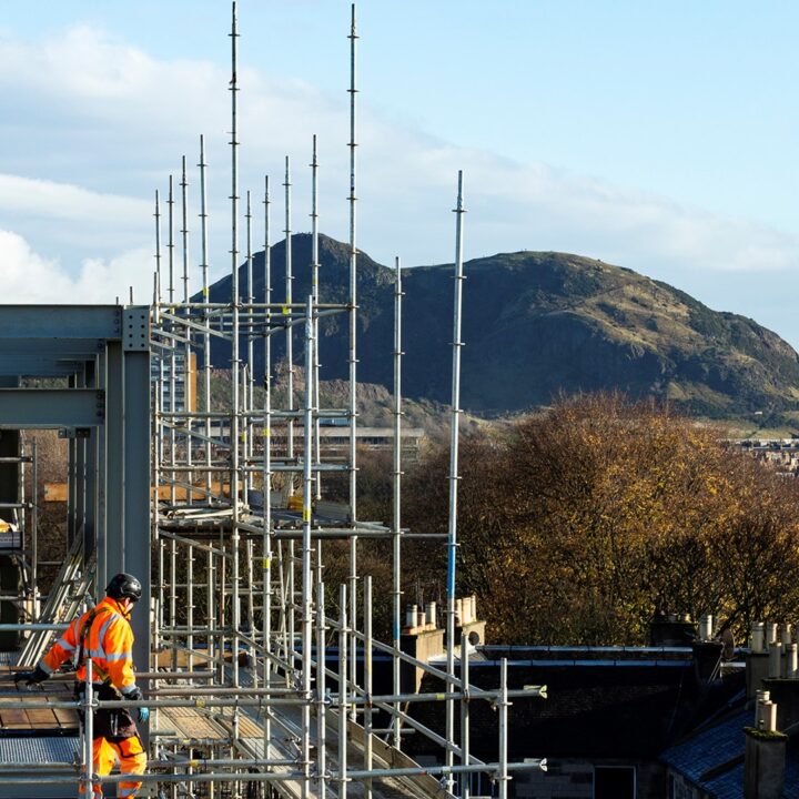 Construction workers in high-visibility clothing work on scaffolding atop a building, with a hilly landscape and blue sky in the background.