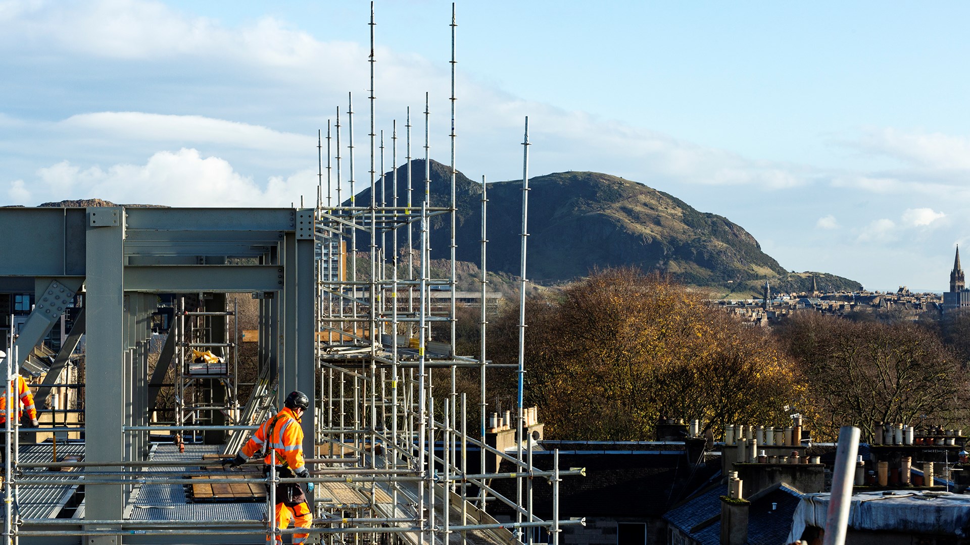 Construction workers in high-visibility clothing work on scaffolding atop a building, with a hilly landscape and blue sky in the background.