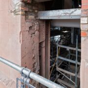 A close-up view of a partially demolished brick and stone wall, showing exposed scaffolding and interior construction materials.
