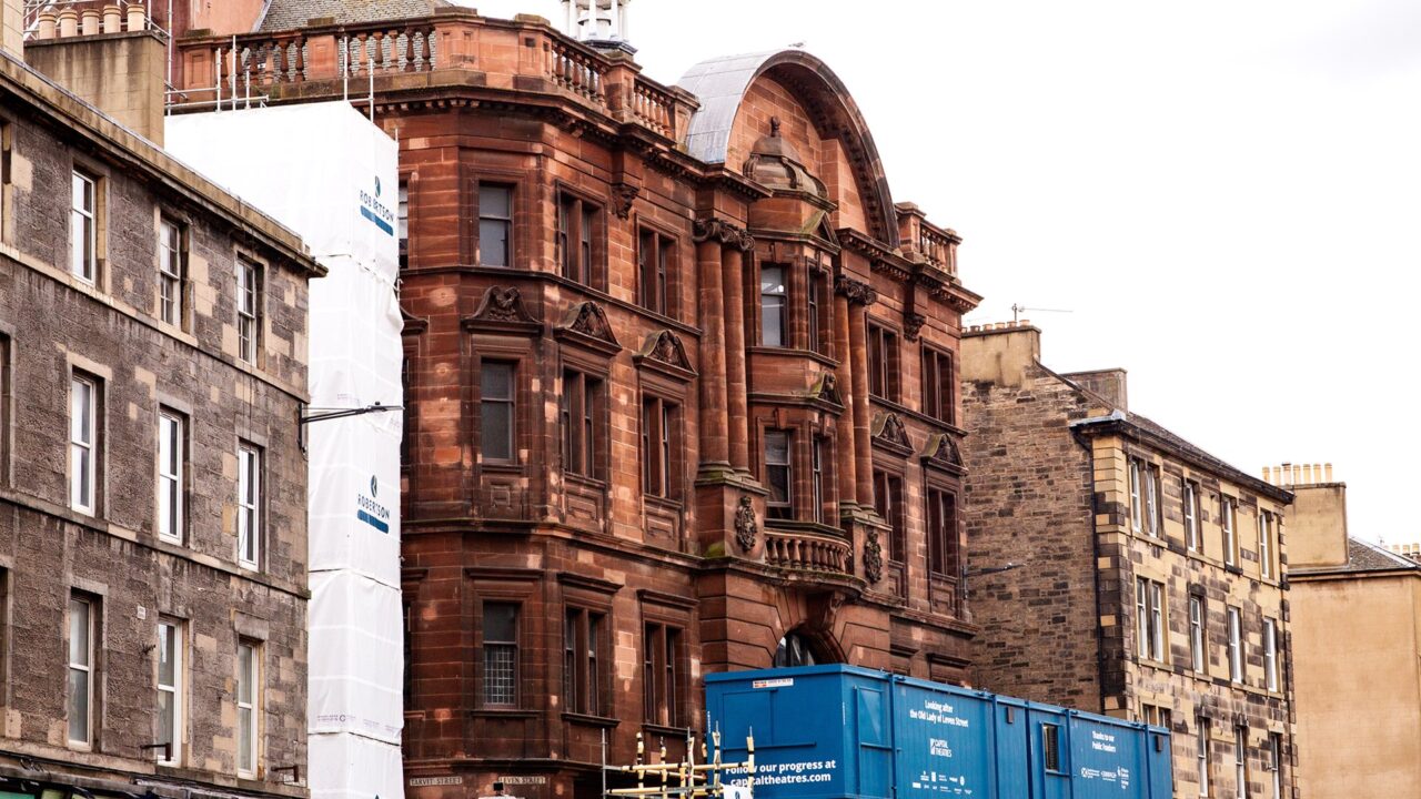 A red sandstone building with ornate architectural details stands between stone buildings, with blue construction containers and scaffolding in front.