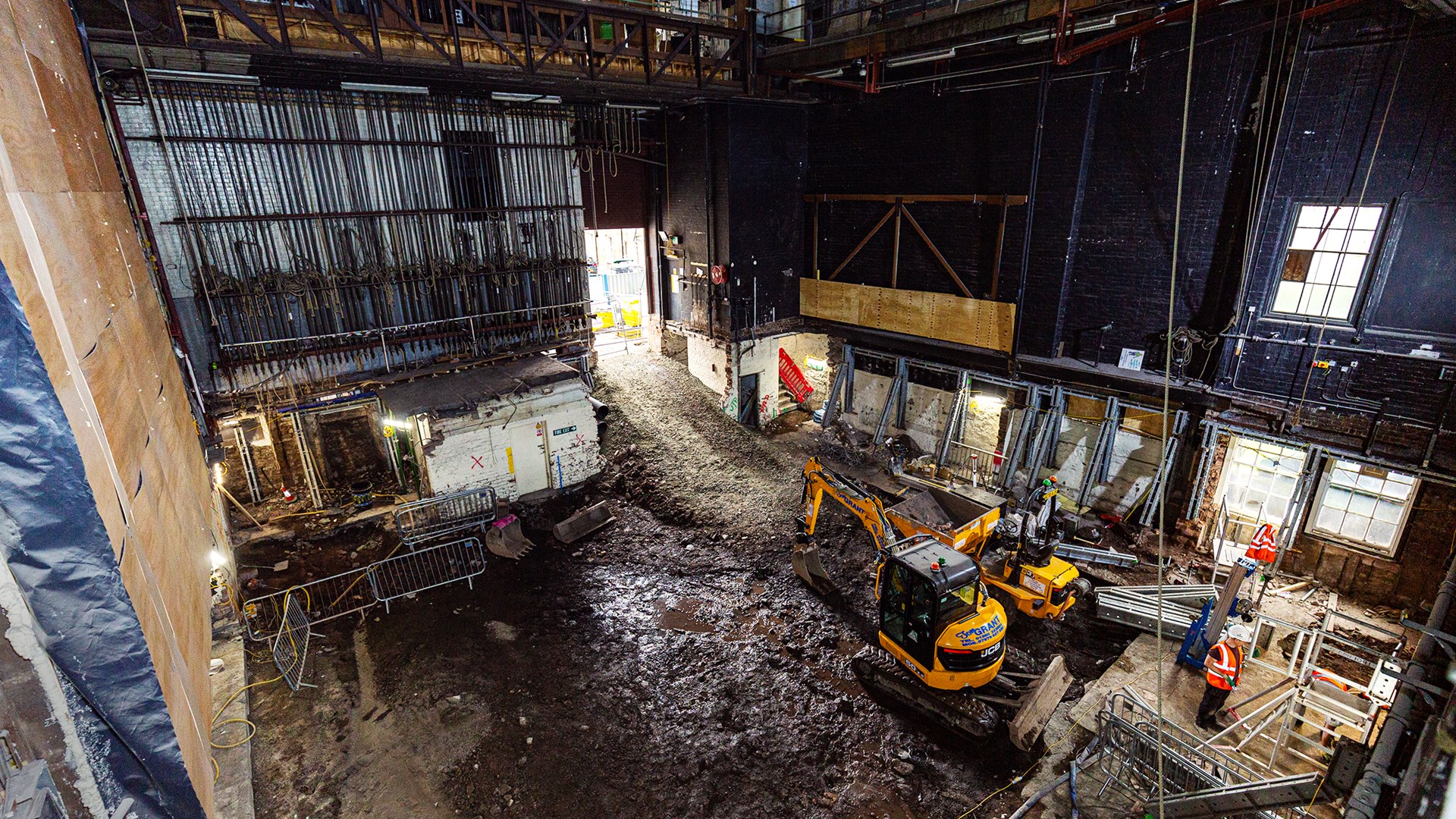 Construction site inside a large building with excavators, mud, barriers, construction materials, and workers wearing safety vests and helmets.