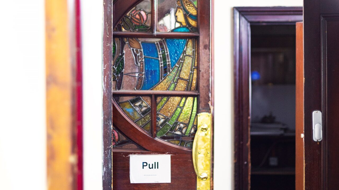 A stained glass door with an image of a person holding a flower has a paper sign reading "Pull" taped to it. The door is partially open, revealing a room beyond.