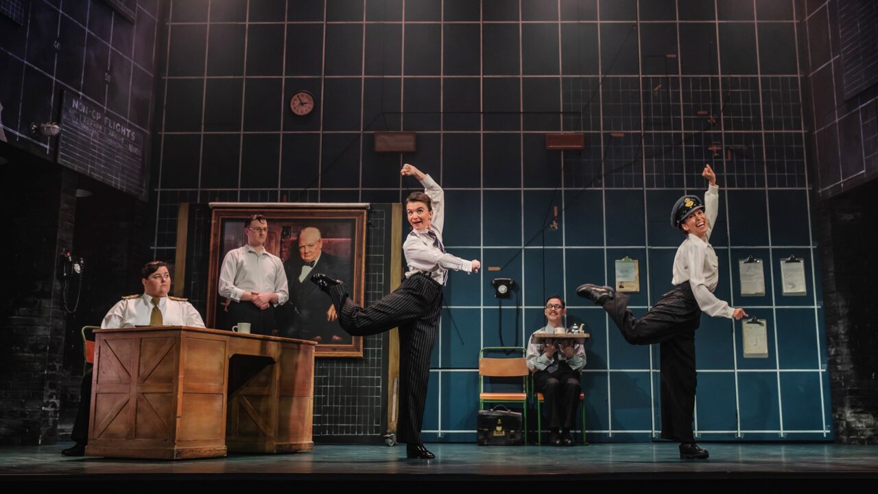 Four actors on stage perform a comedic scene in an office setting, with one actor seated at a desk and the others dancing in mid-step; a large portrait hangs on the wall behind them.
