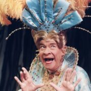 A person in an ornate costume with a large feathered headdress poses with an expressive face and hands raised, wearing a blue and gold embellished outfit.