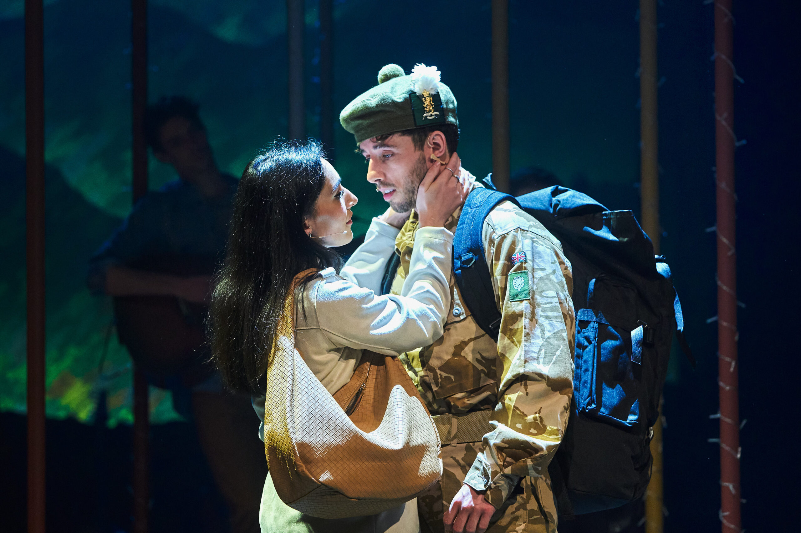 A woman embraces a man in military uniform with a large backpack under stage lighting, suggesting an emotional moment in a theatrical performance.