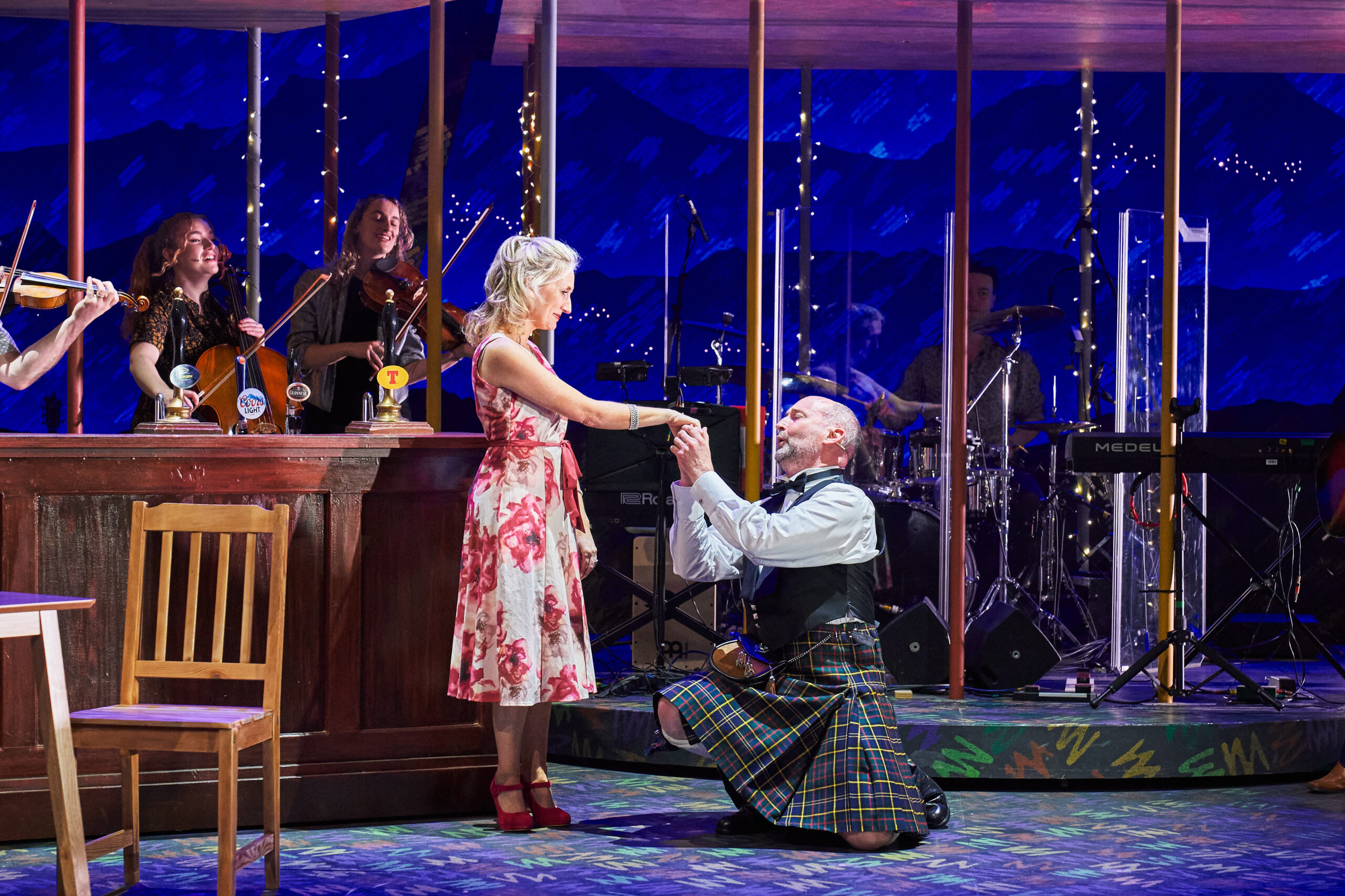 A man in a kilt kneels, holding hands with a woman in a floral dress, onstage beside a bar set, with musicians playing instruments in the background.