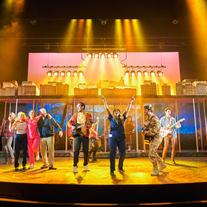 A group of performers sing and play instruments on a brightly lit theater stage with a cityscape backdrop.