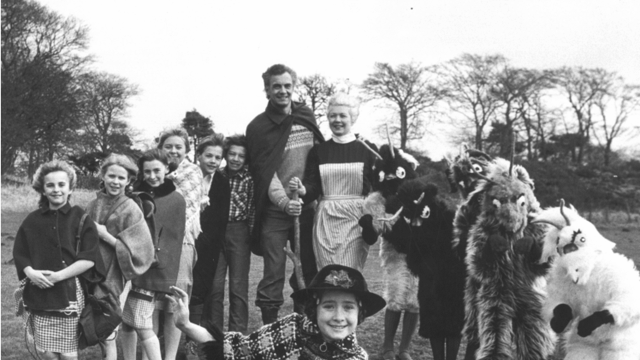 A group of children and two adults stand outdoors, some wearing costumes and animal masks, with trees and open land in the background.