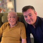 An elderly man in a mustard polo shirt sits in an armchair next to a middle-aged man in a dark shirt; both are looking at the camera indoors.