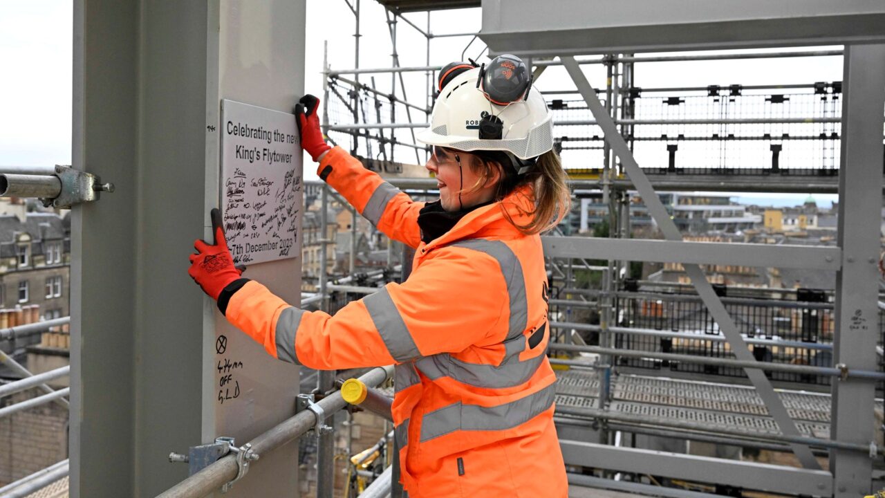 A construction worker in orange safety gear and a white helmet attaches a signed plaque to a metal beam on a building site.