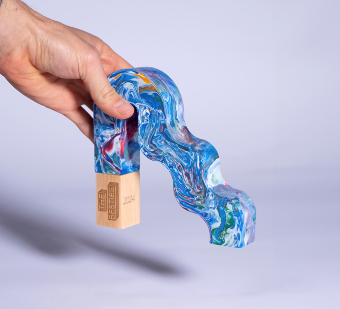 A hand holds a colourful, wavy award made of marbled blue material with a wooden base labelled "THE BESTIES 2024.