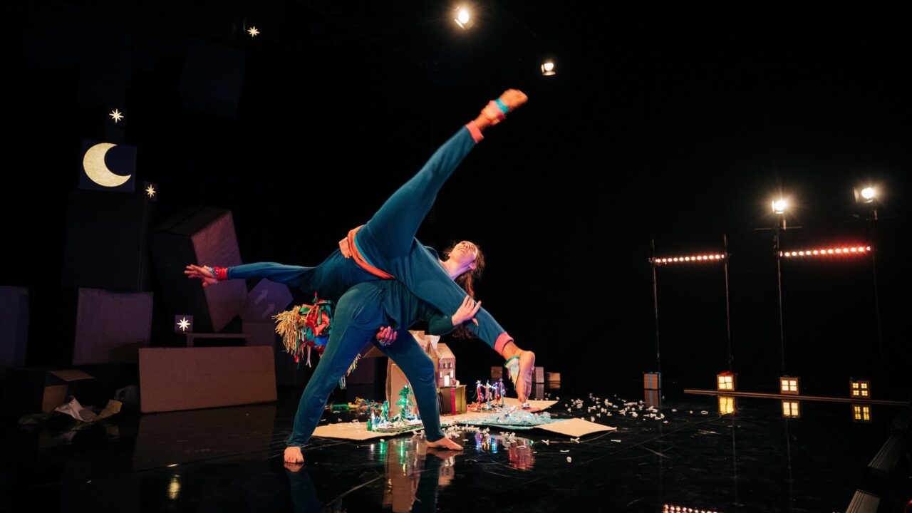 Two dancers perform a balanced acrobatic pose onstage, with one lifting the other. The scene is set in dim lighting with props, a crescent moon, and small lights in the background.