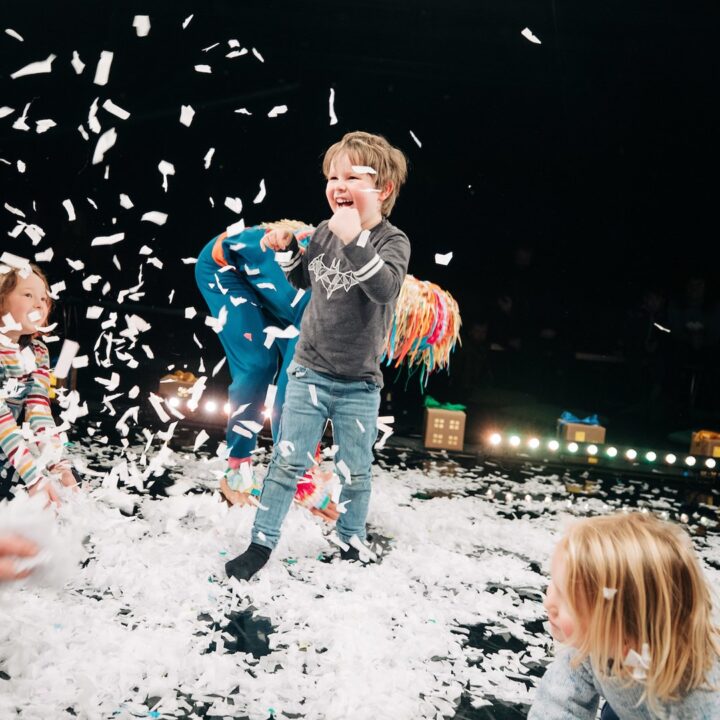 Children play joyfully amid scattered white confetti on a stage, with adults joining in and colorful houses visible in the background.