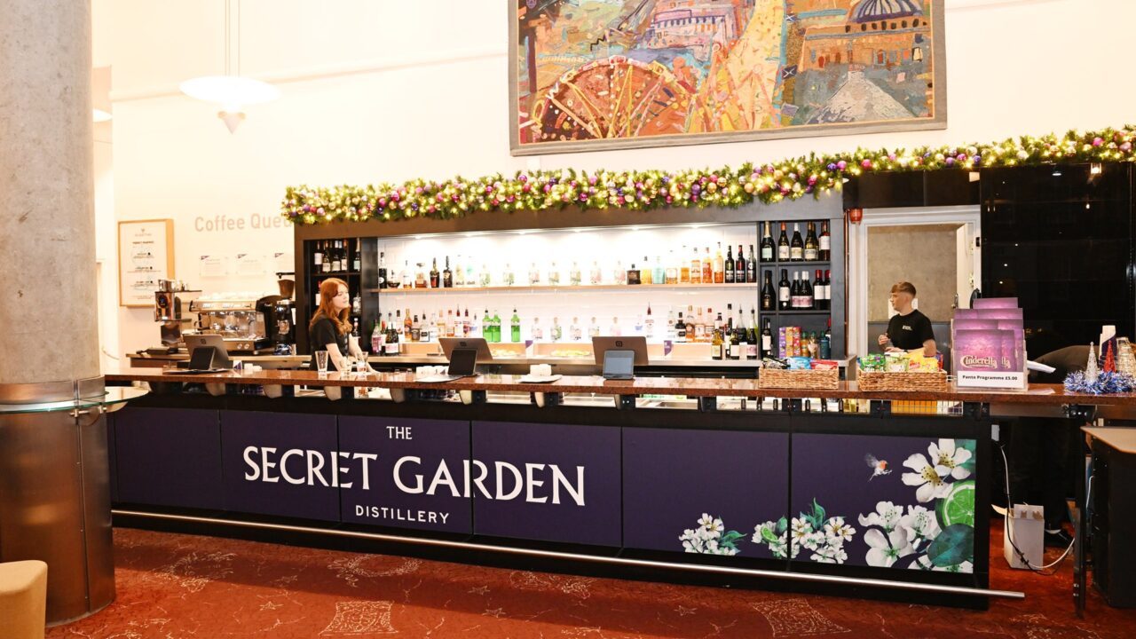 A bar counter labelled "The Secret Garden Distillery" with bottles of alcohol on shelves behind, two staff members, and festive decorations above.