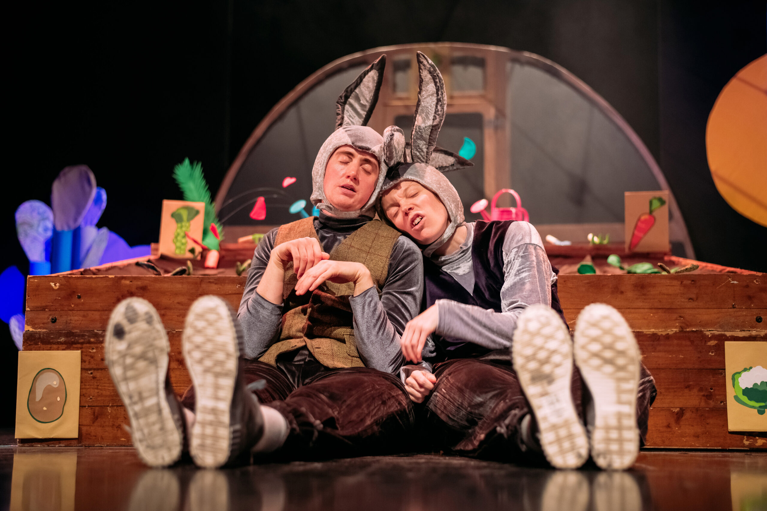 Two people in rabbit costumes sit on the floor, leaning against each other with eyes closed, in front of a colourful stage set featuring vegetables and wooden boxes.